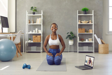 Smiling fit lady doing fitness exercise with workout video lesson at home. Portrait of cheerful positive black woman with happy face expression sitting on yoga mat in living room with laptop computer