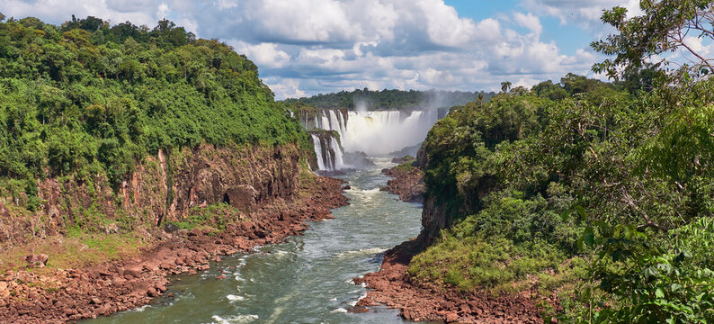Iguazu waterfalls in Argentina, view from Devil's Mouth. Panoramic view of many majestic powerful water cascades with mist. Panoramic image with reflection of blue sky with clouds.