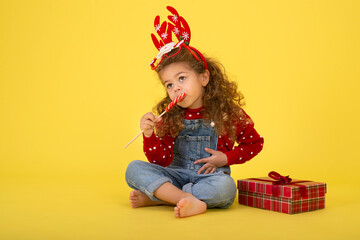 Fototapeta na wymiar Little girl for the Christmas holidays with a gift and a lollipop on a yellow background to copy space