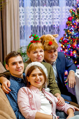 Fototapeta na wymiar New Year's portrait of a family of 4 people wearing carnival hats. Adult children. Christmas tree on the background. Christmas atmosphere. Vertically. Christmas together. Selective focus