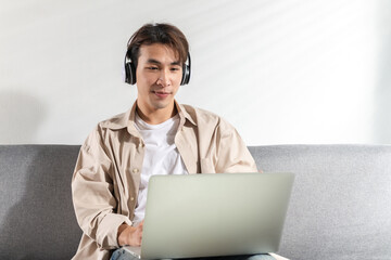 Focused a young smart Asian man wear earphones having video call on a laptop, writing notes in notebook watching webinar video course at home talk using modern technologies and wireless connection