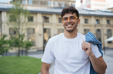 Young smiling Indian man wearing white t shirt, stylish eyeglasses walking on the street. Handsome asian model posing for pictures, looking away