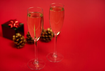 Two flutes of champagne or glasses with sparkling wine on the red background with pine cones ornate with golden paints and luxury Christmas gift in shiny green wrapping paper with glitter red bow