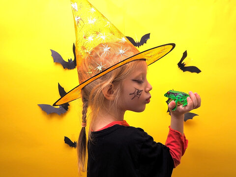 A girl in a witch costume on a background with bats kisses a frog. Child girl dressed as a witch on the background of bats. Preparing for Halloween. Halloween costume.