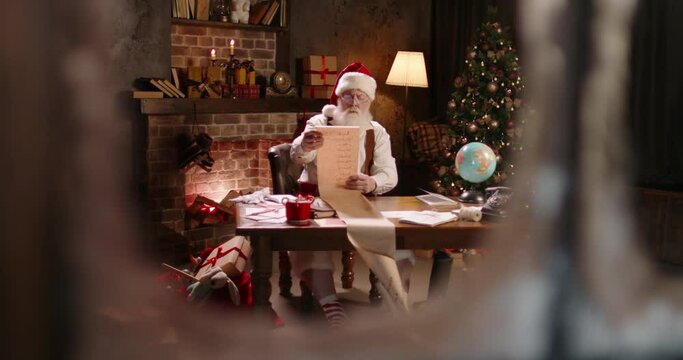 Busy Santa Claus working at night, reading long wishlist, outside view through window. St Nicholas making up checklist of children wishes and presents for kids. Late work in evening on Christmas Eve. - Powered by Adobe