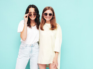 Two young beautiful smiling brunette hipster female in trendy summer clothes. Sexy carefree women posing near blue wall. Positive models having fun. Cheerful and happy in sunglasses