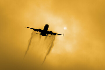 Dark silhouette low-flying commercial plane going to landing leaving contrail against small sun and cloudy sky. Passenger jet arriving at destination on cold day. Concept background in yellow tones