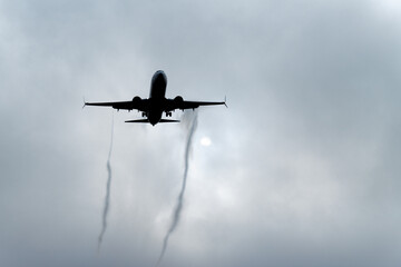 Dark silhouette low-flying commercial plane leaving contrail in cloudy sky going to landing. Air...