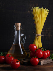 Red ripe cherry tomatoes on a green twig lie on a dark wooden board. Dry pasta spaghetti and olive oil in a jug in the background