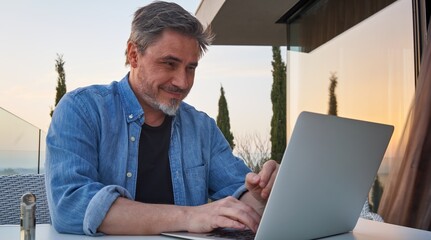 Happy man working online with laptop computer on terrace. Businessman in home office, browsing internet, tele working, video chat.