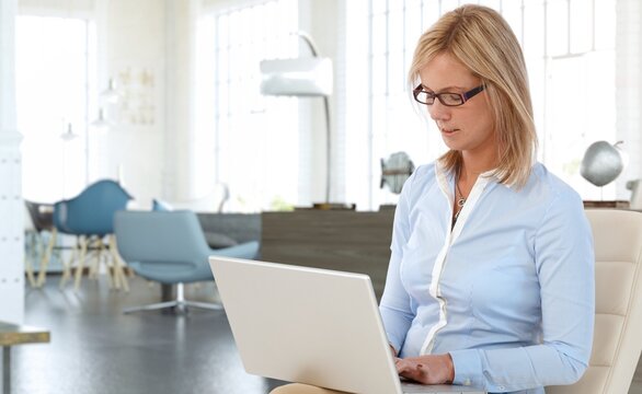 Mid adult woman working online with laptop computer at home sitting at living room. Businesswoman in home office, browsing internet.