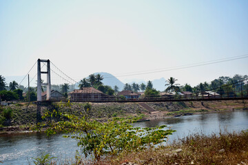 The picture of a suspension bridge across to the village with mountains in the background at noon.
laos country. 2021