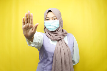 Muslim woman wearing medical mask, with stop sign hand, rejection sign hand, do not keep distance sign hand, do not approach hand sign, isolated on yellow background