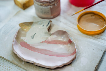 A plate of clay in the shape of a unicorn with a pink mane lies on the table, next to the paint and brush. Creativity in the pottery workshop. - Powered by Adobe