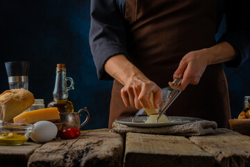 A chef in a dark uniform grates cheese. Lots of ingredients for making Caesar salad, pizza, pie. Wooden texture, blue background. Close-up. Culinary blog, recipe book.