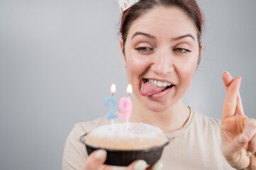 Happy caucasian woman sticking out her tongue and blowing out the candles on the cake with her fingers crossed. The girl celebrates her 29th birthday