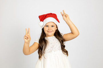 close-up portrait of beautiful long-haired girl in white dress and red Santa Claus hat dancing and showing victory sign with fingers, isolated on white background. 