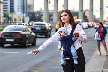 White woman with baby in sling is stopping taxi on traffic street.