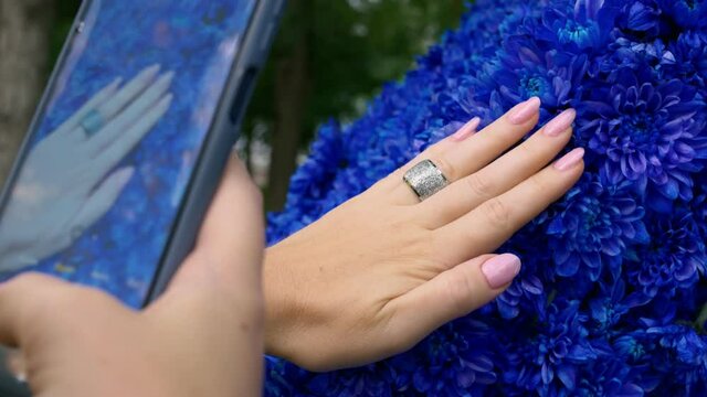 A woman photographs her hand with a beautiful manicure against a background of bright blue flowers. There is a silver ring on the finger. Newest technologies.