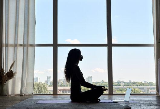 Young calm fit African black woman silhouette sitting on floor at home near window doing yoga breathing exercise, meditating learning online training virtual class on computer. Mental health concept.