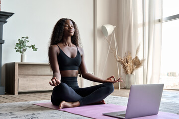 Fototapeta Young calm fit healthy African black woman sitting on floor at home doing yoga breathing exercise, meditating learning online training virtual class on computer. Exercises for mental health concept. obraz