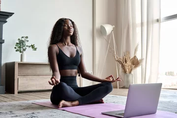 Foto op Canvas Young calm fit healthy African black woman sitting on floor at home doing yoga breathing exercise, meditating learning online training virtual class on computer. Exercises for mental health concept. © insta_photos
