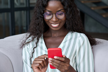 Young happy black African woman wearing glasses using mobile digital apps on cell phone tech device for online shopping or dating sitting on sofa at home, texting on smartphone, writing social post. - obrazy, fototapety, plakaty