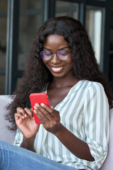 Young happy black African woman wearing glasses using mobile digital apps on cell phone tech device online shopping or dating sitting on couch at home, texting on smartphone, writing social post. - obrazy, fototapety, plakaty