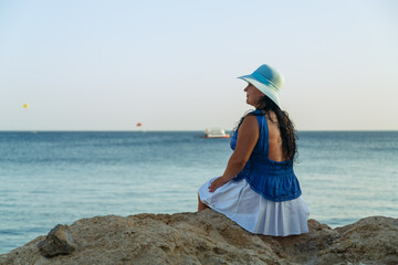 A young brunette woman in a white skirt and blue hat on the seashore sits with her back to the...