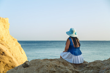 A young brunette woman in a white skirt and blue hat on the seashore sits on the rocks with her back to the camera.