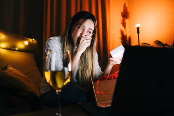 Young millennial woman open gift in front laptop computer during video call and drinking wine, use technology for communicate with friends or family.