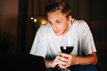 Young millennial man having video call on laptop computer and drinking wine, use technology for communicate with friends or family.