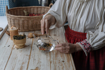 A girl in a folk costume makes tea from herbs and flowers. A woman in the kitchen is preparing her own tea.