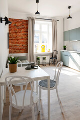 Modern dining room with table and chairs, kitchen, window and bricks wall. Loft apartment in industrial style with minimal design and scandinavian style.