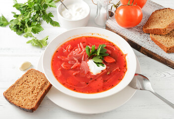 red borscht with sour cream and herbs in a white plate, a spoon, bread on a white table