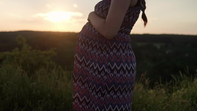 happy pregnant woman touching her tummy on top of mountain in tall grass before sunset. caring mother strokes large belly tummy with hands. pregnancy, motherhood, people, expectation, summer concept