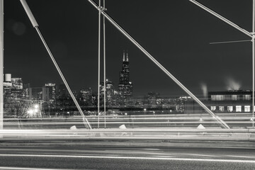 Beautiful black and white night photograph looking through a cable supported bridge towards the Chicago skyline and skyscraper buildings with traffic light trails in the foreground.