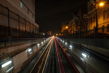 Plakat Long exposure white and red light trails from the CTA trains departing and coming from an underground tunnel at night with alley lights and residential buildings on either side of the tracks.