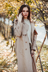 A beautiful tall girl of model appearance in a beige raincoat and gray jeans walks in a city park on a warm autumn day.Lifestyle concept. Autumn street fashion look.