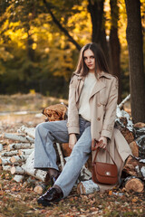 A beautiful tall girl of model appearance in a beige raincoat and gray jeans sits on sawn logs in a city park on a warm autumn day. Autumn street trendy look.