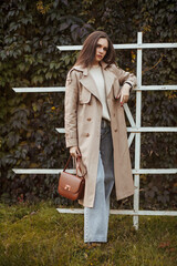 A beautiful tall girl of model appearance in a beige raincoat and gray jeans walks in a city park...