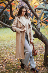 A beautiful tall girl of model appearance in a beige raincoat and gray jeans walks in a city park on a warm autumn day.Lifestyle concept. Autumn street fashion look.