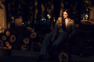 Fototapeta na wymiar A beautiful tall girl of model appearance in a beige raincoat and gray jeans sits on sawn logs in a city park on a warm autumn day. Autumn street trendy look.