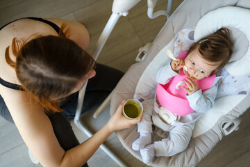 Feeding small caucasian baby wearing silicone bib with broccoli vegetable puree mash concept top view woman mother holding plastic bowl container with food with baby selective focus first meal