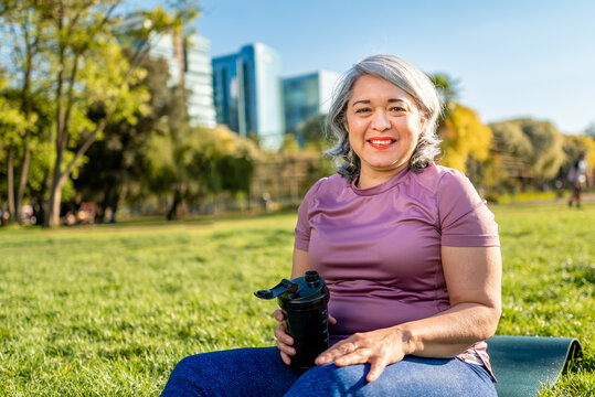 Mature woman smiling and drinking water after a workout exercise at an urban park in the city