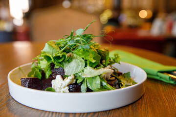 Beetroot salad with arugula, feta cheese and pine nuts in white plate with fork in green napkin over wooden table in the cafe, top view, blurred background