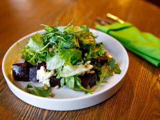 Beetroot salad with arugula, feta cheese and pine nuts in white plate with fork in green napkin over wooden table in the cafe, top view