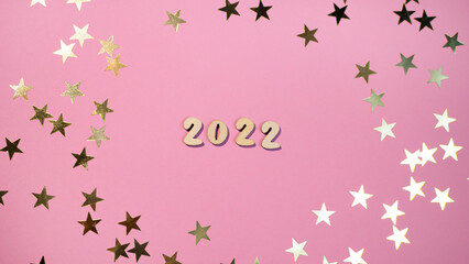 Cheerful wooden lettering happy new year 2022 on a soft gently pink background with gold stars. Sparkle confetti holiday concept 