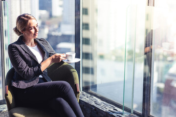 Young business woman holding a cup of coffee. She is wearing white shirt and suit. She is sitting in a hotel lobby.