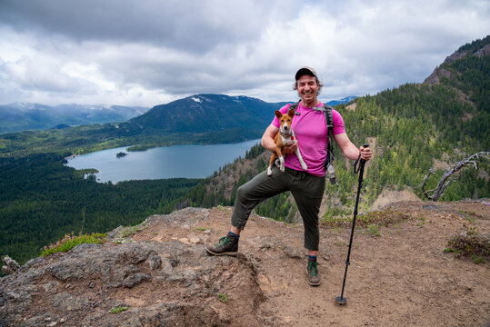 An Athletic Adventurous Male Hiker Standing On Top Of A Mountain With His Dog Looking At The Camera Laughing.